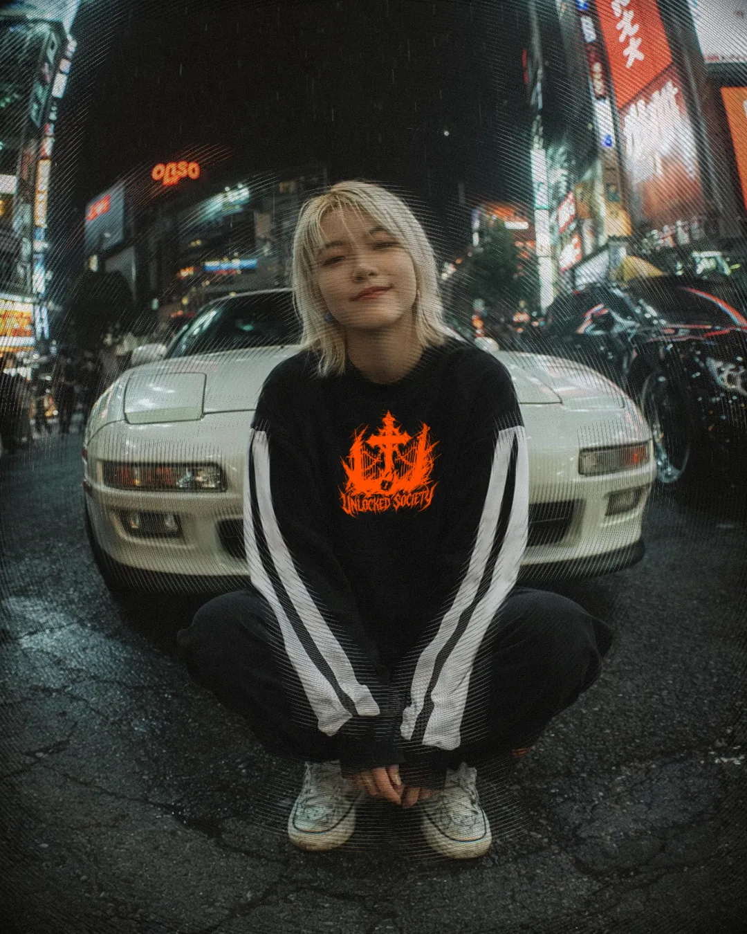 Campagnefoto met fisheye-lens: een persoon in een bedrukt sweatshirt voor een witte sportauto in een neonverlichte straat bij nacht.